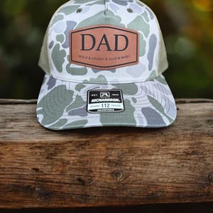 May include: A camouflage-patterned baseball cap with a brown leather patch that reads "DAD." The cap has a green mesh back and a curved brim. The text "HOLLY & LINDSEY & ALLIE & MARLI" is below the word "DAD."