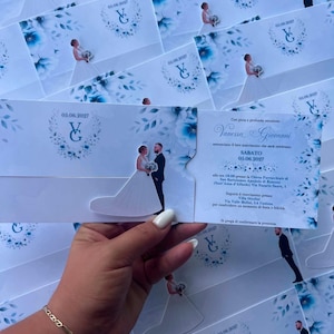Könnte beinhalten: Eine Hochzeitseinladungskarte mit blauem Aquarell-Blumenmuster, einem Monogramm mit den Buchstaben VG und dem Datum 05.06.2027. Die Karte enthält eine ausgestanzte Silhouette eines Paares und gedruckten Text zur Hochzeitszeremonie.