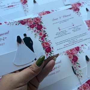 Könnte beinhalten: Eine weiße Hochzeitseinladungskarte mit einer gestanzten Illustration eines Paares in festlicher Kleidung, verziert mit einem Blumenrand aus rosa und dunkelroten Rosen. Der Text enthält die Namen Luna und Clément sowie Details zur Zeremonie.