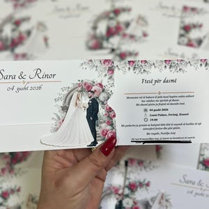 Peut inclure: Une carte d'invitation de mariage avec une illustration découpée d'une mariée et d'un marié devant une arche florale. Le texte indique les noms Sara & Rinor, la date 04 gusht 2026 et les détails de la cérémonie au Leoni Palace à Ferizaj, Kosovë.