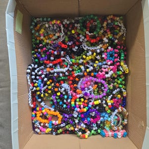 May include: A cardboard box filled with colorful beaded bracelets. The bracelets are made with small, colorful beads and have letters spelled out on them.