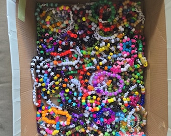 Può includere: Una scatola di cartone piena di braccialetti di perline colorate. I braccialetti sono fatti con piccole perline colorate e hanno lettere scritte su di loro.