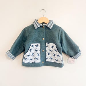 Peut inclure: Une veste sarcelle avec un col et des revers de manches à carreaux vichy bleus et blancs. La veste présente deux poches avant avec un motif en tissu blanc représentant des crabes bleus. Elle possède trois boutons-pression métalliques.