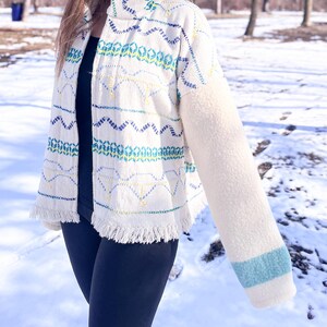 May include: A cream-coloured jacket featuring a high collar and a front zip closure. The body displays horizontal rows of embroidered geometric patterns in blue, teal, and yellow. One sleeve is solid cream fleece with a teal stripe at the cuff.