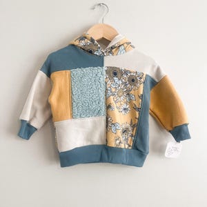 May include: A long-sleeve hooded sweatshirt featuring a patchwork design of blue waffle-knit fabric, solid mustard yellow panels, cream-coloured sections, a textured light blue sherpa square, and a floral print with sunflowers on a mustard background.