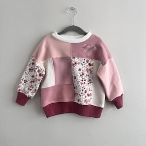 May include: A patchwork sweatshirt in pink, cream, and burgundy, with floral and gingham patterns. A small heart-shaped patch is sewn on the upper right. The cuffs and hem are burgundy. The sweatshirt is hanging on a grey hanger.