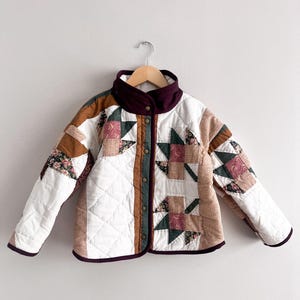 Peut inclure: Veste matelassée avec un motif patchwork. La veste présente une base blanche avec des coutures en losange, rehaussée de carrés marron, verts et à motifs floraux. Elle a un col et une bordure bordeaux, et se ferme par des boutons sur le devant.