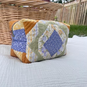 Peut inclure: Une trousse rectangulaire zippée fabriquée en tissu matelassé avec un motif patchwork. L'extérieur présente une combinaison de motifs floraux et rayés dans des tons jaune moutarde, vert sauge et bleu pervenche avec un losange central.