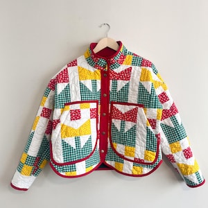 May include: A quilted jacket with a patchwork design in red, yellow, green, and white. The jacket has a red collar, pocket trim, and lining. It features a button-up front and two front pockets. The jacket is hanging on a wooden hanger.