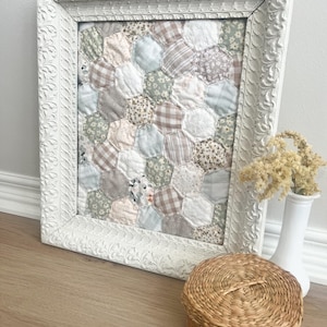 May include: A framed textile artwork with a patchwork design of hexagonal fabric pieces in various patterns and colours, including floral, gingham, and solid fabrics. The frame is white with ornate detailing.