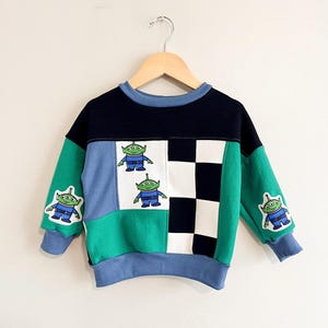 May include: A long-sleeve sweatshirt featuring a colour-blocked design in navy blue, light blue, and green. The front panel displays a white section with two green three-eyed alien characters and a black and white checkerboard pattern.