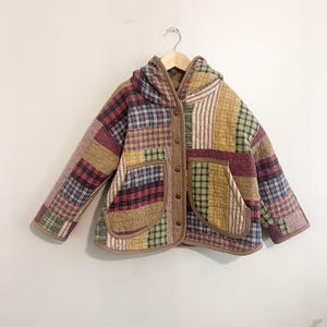 Peut inclure: Une veste matelassée présentant un motif patchwork composé de divers carrés de tissu à carreaux et unis dans des tons de rouge, jaune, vert et bleu. La veste comprend une capuche, des manches longues, deux poches avant et un liseré marron.