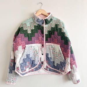 Könnte beinhalten: Gesteppte Jacke mit Patchwork-Design. Sie hat einen hohen Kragen, einen Knopfverschluss und zwei Vordertaschen. Der Stoffmix besteht aus floralen und geometrischen Mustern in Rosa, Grün, Blau und Weiß, mit rosa Einfassungen.
