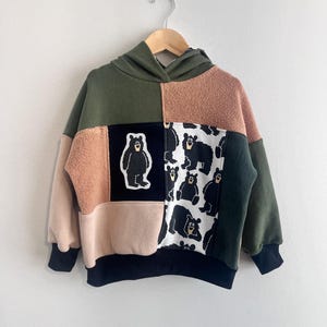 May include: A colour-blocked hooded sweatshirt featuring panels of olive green, tan, and black fabric. The front displays a central black patch with a single bear illustration and a side panel featuring a repeating pattern of black bears on a white background.