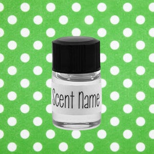 May include: A small, clear glass bottle with a black cap. A white label with the words "Scent Name" in black script is affixed to the bottle. The background is a bright green fabric with white polka dots.