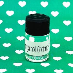 May include: A small clear glass bottle with a black lid and a white label that reads "Bergamot Coriander". The bottle is sitting on a green background with white hearts.