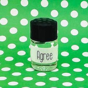 May include: A small, clear glass bottle with a black cap. The bottle has a white label with the word "Agree" in black script. The background is a bright green with white polka dots.