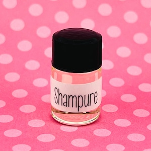 May include: A small clear glass bottle with a black cap and a white label that says "Shampure". The bottle is sitting on a pink background with white polka dots.