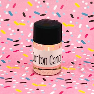 May include: A small clear glass bottle with a black lid and a white label that says "Cotton Candy". The bottle is sitting on a pink and white background with colorful sprinkles.