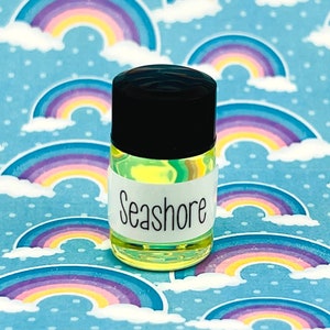 May include: A small, clear glass bottle with a black lid and a white label that reads "Seashore". The bottle is filled with a yellow liquid and is sitting on a blue and white background with rainbow patterns.