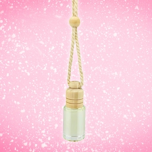 May include: A small, clear glass bottle with a wooden cap and a beige rope for hanging. The bottle is filled with a light-colored liquid. The background is a pink gradient with white speckles. This item is likely an air freshener or perfume.
