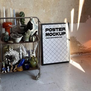 May include: A black-framed rectangular poster mockup featuring a white background with a black diamond grid pattern and the bold black text POSTER MOCKUP PREMIUM FRAME MOCKUPS. Beside it stands a two-tiered metal rolling cart filled with various ceramic vases, bowls, and twisted candles.