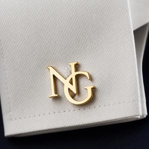 May include: Gold-colored cufflinks with the intertwined letters "NG" are displayed against a white dress shirt cuff. The cufflinks have a polished finish, reflecting light. The shirt cuff is made of a textured, white fabric.
