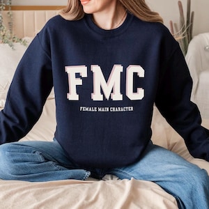 May include: Navy blue sweatshirt with the letters "FMC" in white block letters, with the words "FEMALE MAIN CHARACTER" below. The sweatshirt is worn with blue jeans.