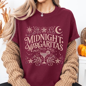 May include: Burgundy t-shirt featuring the text "MIDNIGHT MARGARITAS PRACTICAL MAGIC" in an ornate font. The design includes stars, a crescent moon, and a margarita glass. The t-shirt is a crew neck style.