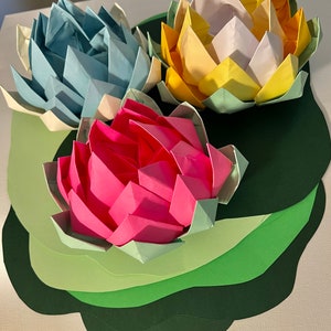Op de afbeelding: Drie origami-lotusebloemen in blauw, roze en geel, gerangschikt op een groene gelaagde papieren achtergrond.