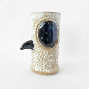 May include: A handmade ceramic mug with a unique owl design. The mug features a textured, off-white body with a dark blue eye and a black beak. The mug is approximately 5 inches tall.