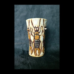 May include: A cylindrical ceramic cup with a cream-colored glaze featuring a repeating relief pattern of brown, insect-like figures with segmented bodies and antennae. The figures are arranged vertically around the exterior of the vessel.