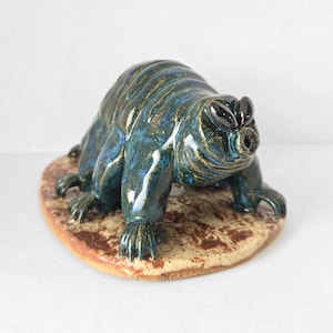 May include: A ceramic sculpture of a stylized animal with a blue and gold glaze. The creature has a rounded body, small legs, and a pig-like snout. It rests on a brown, oval-shaped base, creating a unique decorative piece.