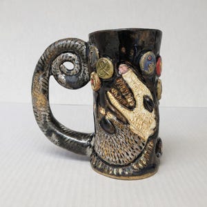 May include: A handcrafted ceramic mug with a unique design. The mug features a dark, textured exterior with a sculpted handle and decorative elements. The design includes a stylized animal face and colorful accents, creating a rustic and artistic aesthetic.