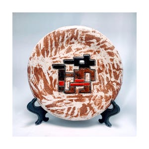 May include: A round ceramic plate with a textured, off-white and brown surface. The plate features a geometric design in black, brown, and red. The plate is displayed on a black metal stand.