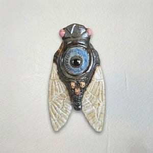 May include: A ceramic cicada sculpture featuring a dark brown, glossy body with a prominent central blue eye. The insect has two light beige, textured wings with fine dark speckles and two small pink spheres positioned at the top of the head.