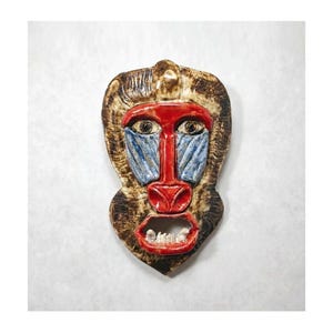 May include: A ceramic baboon mask with a detailed, hand-painted design. The mask features a red face and mouth, blue facial markings, and brown fur-like details around the edges. The baboon's mouth is open, revealing white teeth.