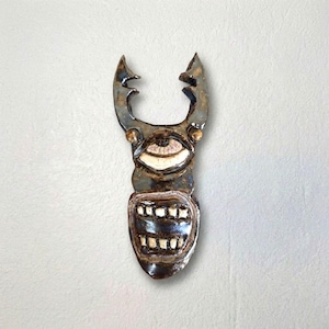 May include: A dark brown and metallic glazed ceramic wall sculpture shaped like a stylized creature with two upward-curving horns, a single large central eye with a white iris, and a wide mouth displaying two rows of square white teeth.