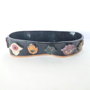 May include: An oval-shaped black ceramic bowl with a glossy finish. The exterior features a series of small, raised hamsa hand symbols in various colors including white, orange, green, and purple, each centered with a small dark circular detail.