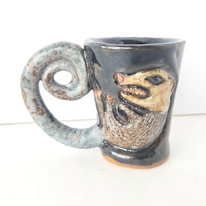 May include: Handmade ceramic mug with a unique, spiraled handle and a textured possum design. The mug is primarily dark blue with brown and tan accents, and a light blue handle. The possum has a pink nose.