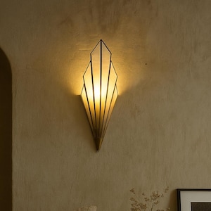 May include: An Art Deco-inspired wall light with a geometric design, made of gold-toned metal. The light emits a warm glow, mounted on a textured, beige wall. A sofa with a brown cushion and throw is in the foreground. The room has a neutral colour palette.