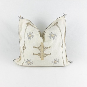 May include: Cream-colored decorative pillow with embroidered geometric patterns in shades of brown and gray. The pillow has a textured appearance and a dark brown stitched border. The design features star-like and linear motifs.