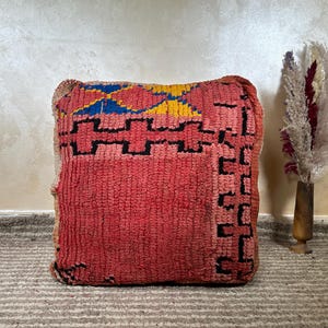 May include: A square, red Moroccan pouf with a geometric design in black, yellow, and blue. The pouf has a textured, woven appearance and is likely made of wool. The design is a traditional Berber pattern.