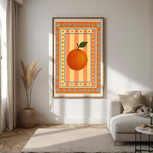 May include: A framed art print featuring a large orange fruit with a green leaf, set against a striped background of orange and yellow. The artwork is framed in a wooden frame and has a decorative border. The print is displayed in a living room setting.