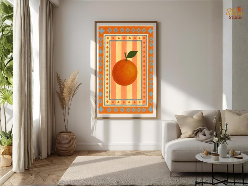 May include: A framed art print featuring a large orange fruit with a green leaf, set against a striped background of orange and yellow. The artwork is framed in a wooden frame and has a decorative border. The print is displayed in a living room setting.