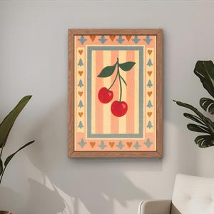 May include: A framed art print featuring a pair of red cherries with green leaves against a striped background. The artwork is in a wooden frame and has a decorative border with hearts and floral elements. The print is hanging on a gray wall.