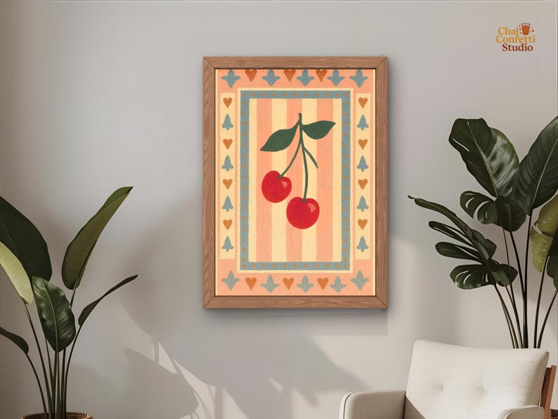 May include: A framed art print featuring a pair of red cherries with green leaves against a striped background. The artwork is in a wooden frame and has a decorative border with hearts and floral elements. The print is hanging on a gray wall.