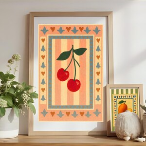 May include: Framed art prints featuring a cherry and an orange design. The cherry print has a red and green color scheme, while the orange print has a yellow and green color scheme. The prints are displayed on a wooden surface with plants.