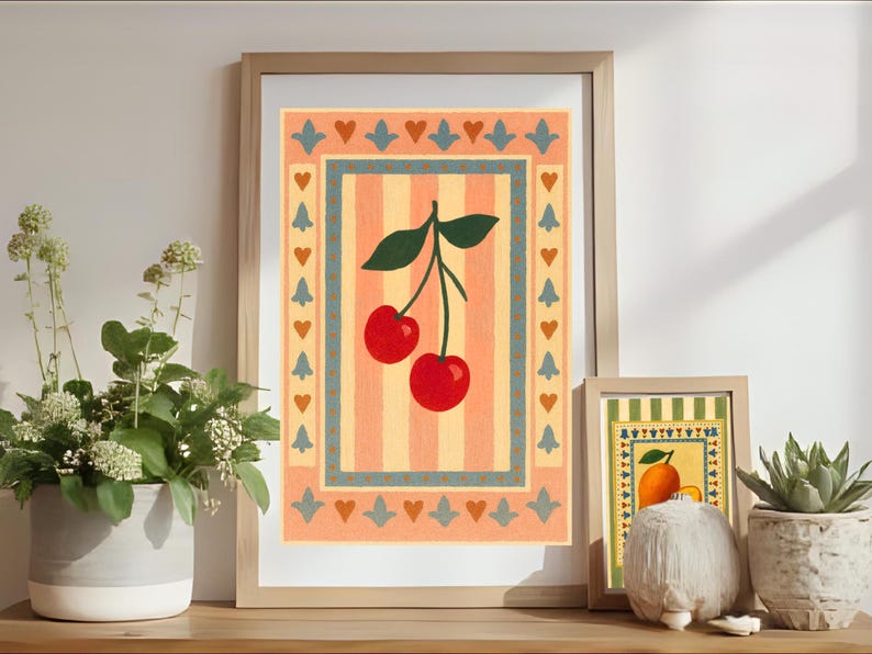 May include: Framed art prints featuring a cherry and an orange design. The cherry print has a red and green color scheme, while the orange print has a yellow and green color scheme. The prints are displayed on a wooden surface with plants.