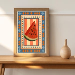 May include: A framed print featuring a watercolor illustration of a watermelon slice. The artwork has a red, orange, and blue border with a striped background. The print is displayed on a wooden table.